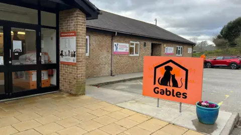 The Gables Cats and Dogs Home entrance. The door is on the right with a black frames and windows. On the right is a vibrant orange sign with a silhouette of a dog and a cat under a roof, with the word 'Gables' below. There is a blue plant pot beside it and a car parked in the parking area behind