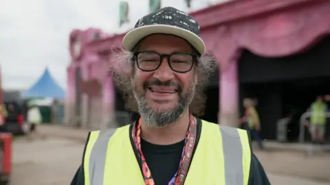 Natty looking direct to camera and smiling. He has on a black and white hat and black rimmed glasses. He is wearing a high-vis vest.