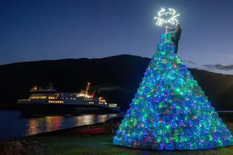 Jane Barlow/PA Media Gordon Wink places the "star" on to top of the village's creel tree. The star is the outline of crab decorated with white lights. In the sea nearby is CalMac's ferry MV Loch Seaforth. It is dark and the lights on the tree and the ferry are on.