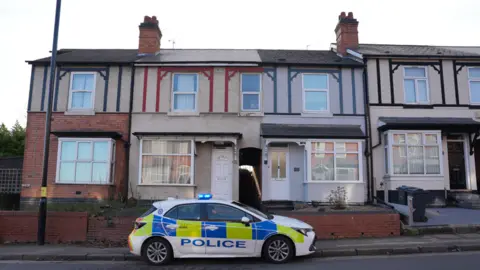 BBC A police car parked outside the penultimate house at the end of a row of terraced houses. The red-brick and rendered houses have two stories and timbers painted in black, red and blue.