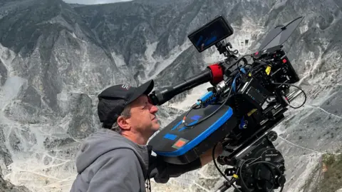 A24 Lol Crawley, who has short grey hair and is wearing a black baseball cap and grey hoody, looking through the lese of a large black camera which is pointed upwards, at something off to the right of the picture. In the background are huge grey mountains.