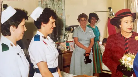 North West Anglia NHS Foundation Trust  Nurse Mary Donaldson pictured in the hospital wearing a white nursing uniform which has a blue epaulette on the shoulder and a matching blue belt around her waist. She has short brown hair, a white hat and is smiling at the late queen Elizabeth. The late Queen holds flowers on a visit to the ward and wears a red and black checked blazer with a matching hat. 