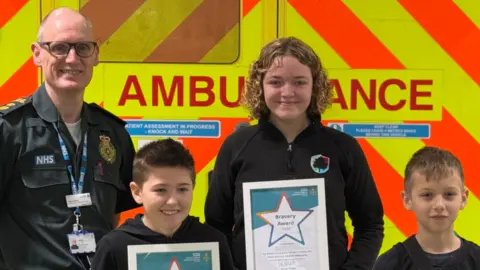 East of England Ambulance Service Simon Chase, a paramedic, is standing in a row next to three children. They are looking directly at the camera and smiling. The children are each holding a certificate. Behind the group is the back of an ambulance.