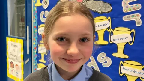 Elena is looking directly at the camera. She has blond hair tied back in a ponytail. She is wearing a light blue shirt and jumper. She is clearly in a school as the background has a wall display featuring paper trophies and names.