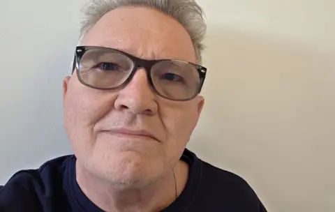 Kevin Jones A close up selfie of Kevin Jones shows him looking straight at the camera wearing glasses and a black T Shirt against a neutral beige wall.