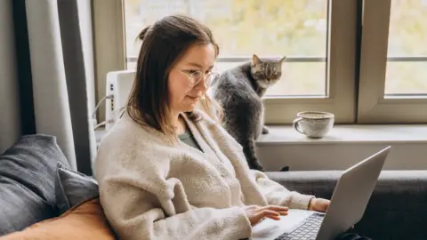 Getty Images A young woman working from home on a laptop, sitting on a sofa and watched by a grey cat on the window sill. 