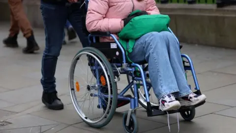 A person is being pushed in a wheelchair. We can't see anyone's face. The person in the wheelchair is wearing blue jeans and a pink jacket. It looks like one of her trainers has become untied. Pushing them, is a person wearing dark shoes and dark jeans.