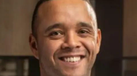 BBC Michael Phipps, who is black and has short dark hair, is smiling for the camera. 