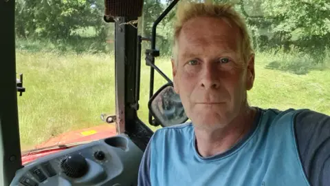Richard looks into the camera with a black face. He's wearing a light blue t-shirt and is sitting in a tractor. Green grass can be seen in the background.
