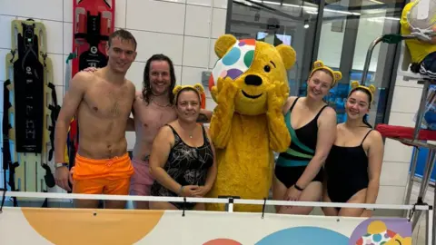 Lauren Carter/BBC Pudsey bear is in the middle of the group of swimmers, they are smiling at the camera, three are wearing Pudsey ears