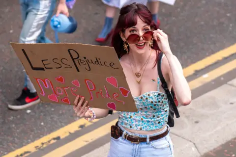 PA Media A participant in a Pride parade looks up toward the camera while holding a handmade cardboard sign reading “Less Prejudice More Pride,” decorated with pink hearts. The participant wears large tinted sunglasses, a sequined top, and light blue jeans while standing on a city street.