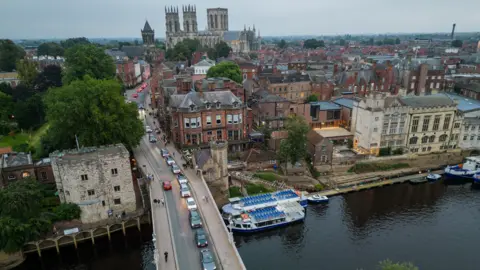 An aerial shot of York. A long line of traffic is queuing to cross Lendal Bridge at the bottom of the frame over the River. In the distance York Minster can be seen towering above the city.