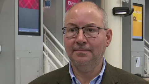 A man wearing a suit and shirt standing in front of check-in desks at Jersey Airport.
