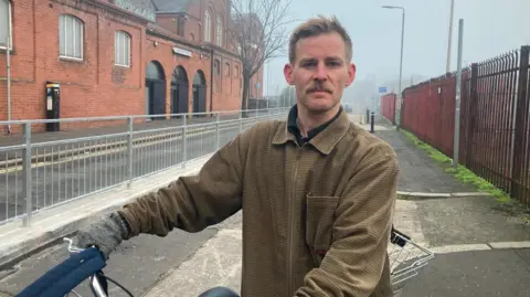 A man wearing a brown above the shirt stands in a bike lane, which puts his bicycle handber aside. There is a basket behind the bike. He has fair hair and brown mustache. 