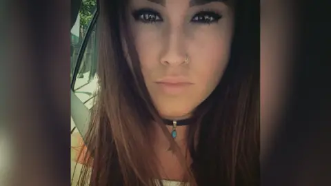 A selfie of a young woman with long dark hair. She is wearing heavy eye makeup and a nose ring and has a black chocker necklace around her neck. She appears to be in a car, with the seat belt visible.