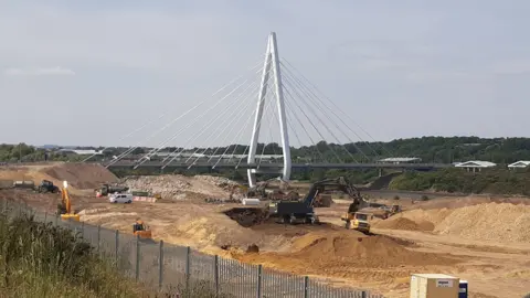 The construction site of the Crown Works film studio. A large area of land is dug out. There are diggers dotted around the site. A white bridge borders is on one side of the site.