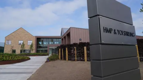 A general view of HMP and YOI Stirling shows a modern, blonde-bricked two-story building with new landscaping surrounding it and a sign with " HMP and YOI Stirling " in large black letters.