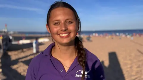 A young woman on the beach, with the sand and sea behind her and a blue sky. She is wearing a purple polo shirt and her brown hair is braided. 