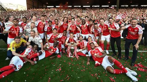 Getty The Arsenal men's squad and coaching staff pose for a team photograph on the pitch with the Premier League trophy, surrounded by red and white confetti, with fans visible in the stands behind them.