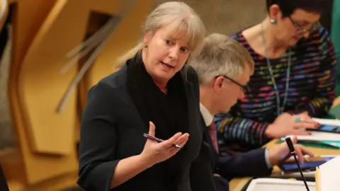 PA Media Shona Robison, a woman with blonde hair, speaks in the Scottish Parliament chamber with her right arm, bent at the elbow, held in front of her. She is holding a pen. She is wearing a black top and is visible from the waist up.  