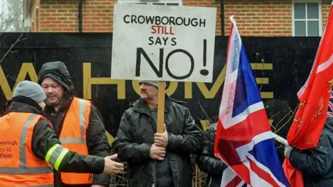 A man in a black jacket is holding a sign saying Crowborough Still Says No, with two people in orange hi vis vests on his left, and people carrying flags to his right