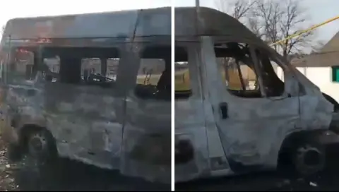 Tetiana Rudnik An ambulance on which any paintwork has been burned off by shelling. There are signs of scorching across the plain white body. Glass has gone from the windows and there appears to be little left of the inside of the vehicle.
There is a bare tree in the background.