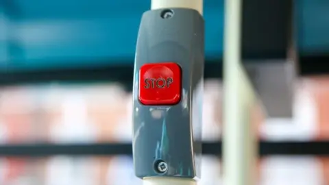 Getty Images A stop button on a bus. The button is red and the casing around it is blue. It is attached to a pole and has the word stop in capital letters embossed into it.