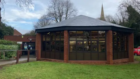 A side view of Octagonal brick building with a large windows. It has a pointed slate roof and grass in front.