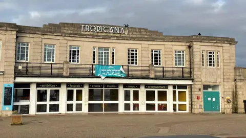 Old seaside building on the seafront. It is a classic opening to an old lido on a road. There is a big sign at the top that says "Tropicana" with a cloudy, moody sky.
