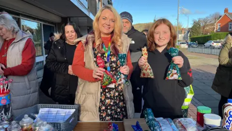 A blonde woman holding Christmas-decorated sweets is stood next to a child with ginger hair. They are also holding a bag of sweets in each hand as they stand in front of a tuck shop with various treats and drinks.