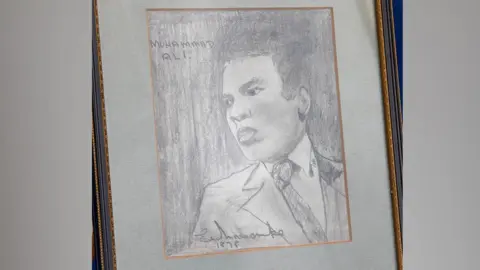 Hansons Auctioneers A black and white sketch of Muhammad Ali by Eric Morecambe is seen mounted on an easel. It shows a depiction of Ali wearing a suit and tie and is signed by Morecambe
