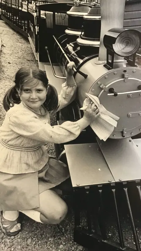 A young woman smiles at the camera as she kneels by a miniature railway in a black and white image