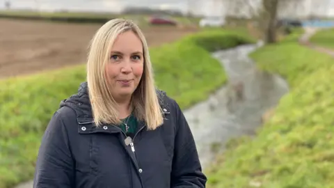 A woman with blonde hair and a blue coat is standing in front of a fast flowing stream