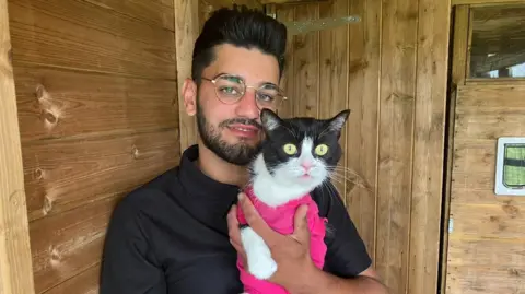 A man in a black t-shirt, with black hair and a short black beard and glasses is sitting in a wooden cabin. He is holding a three-legged black and white cat wearing pink protective clothing.