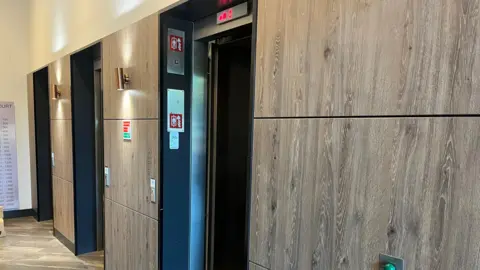 An indoor corridor with wood‑panelled walls and three lift doors, one of which is open, with red digital indicators above and safety signage mounted beside the lifts.