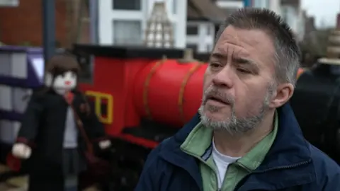 Leon Girling gives a TV interview in front of his display, including a large model of a train. He has a grey beard and dark spiky hair and wears three tops including a dark fleece.