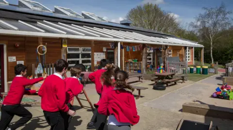Getty Children in red school uniform jumpers and dark trousers run towards a school building.