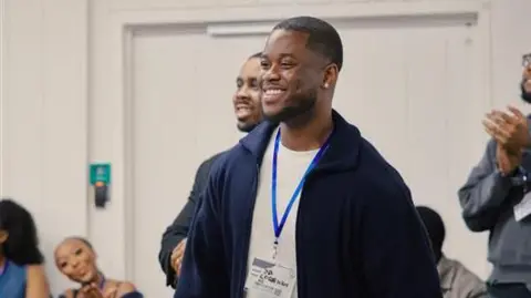 Michaela Ajani/M.ShotIt Jordan Boateng is picture standing in the distance looking away from the camera and smiling. He's a black man with short, dark hair and is wearing dark trousers, a white t-shirt and a navy jacket. He's leaning on a table with his left hand and there's other people in the background who also appear to be smiling and clapping while looking at the same thing Jordan is.