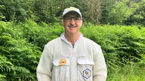 A man with spectacles and a baseball cap, wearing a white beekeeping suit, stood in front of a wooded area