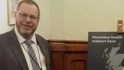 Nobby Clarke Nobby Clarke smiling at the camera in a grey suit jacket and white shirt and striped purple tie. On the right is a board that says 'Homeless health matters here' in white text.