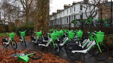 Reuters A park in autumn with orange leaves on the ground and around a dozen green hire bikes clustered together. One bike is on its side on the ground. In the background is a row of white Victorian houses and more green hire bikes on the pavement.  