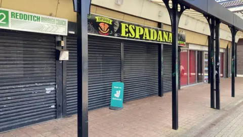 An empty row of shops in Woodrow, in Redditch - the image shows some old retail units protected by brown metal shutters.