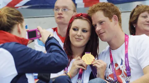 Getty Images Jessica-Jane Applegate having her picture taken with Prince Harry while they are sitting next to each other. Applegate is showing her gold medal to the camera while someone with their back to the camera is taking a picture of the two of them.