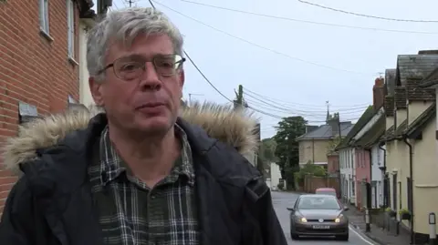 A man stands near a road in a village on a grey day. He has short grey hair and sunglasses and is wearing a chequered green shirt with a parka coat over the top. There is a grey car on the road travelling toward the camera. The road is lined with homes.