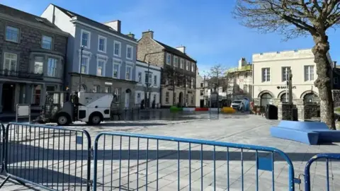 BBC Market Square, a large area with grey tiling, there are stone buildings in the background, and a blue barrier in the front, there is a street cleaner vehicle, and no othervehicles in the space.