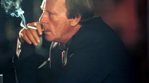 Dennis Waterman: A man with brown hair looking to his right and wearing a dark jacket. He is hold a white cigarette with a brown tip to his lips with his right hand. Smoke is issuing from the cigarette.