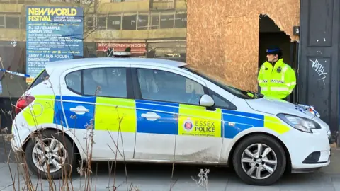 A female police officer wears a hi-vis jacket and black hat as she guards a vacant shop, which has been partially boarded up. She stands behind a hatchback police car and next to blue and white tape.
