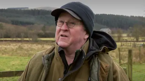 A man in a black hat with glasses and a green jacket stands in front of a fence with tree-lined hills in the background