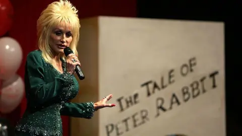 Dolly Parton, holding a microphone and stood in front of a giant book, is stood next to a giant sized book which says The Tale of Peter Rabbit.