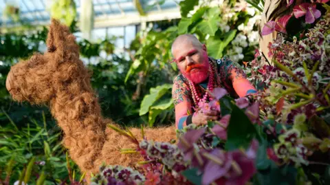 PA Media Man with pink facial hair tends to some flowers of the same hue.